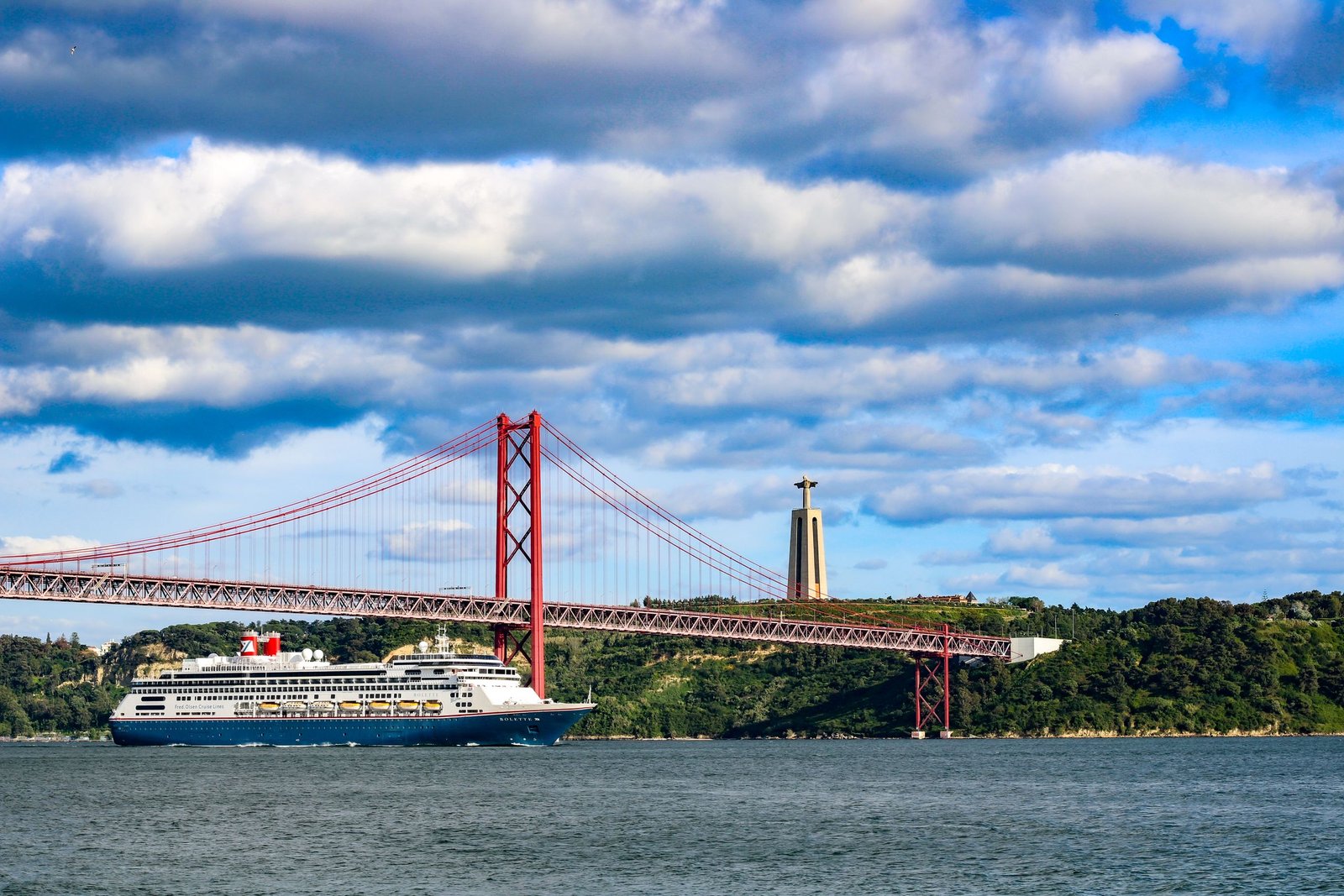 Scenic view of a Fred Olsen cruise ship sailing under the iconic 25 de Abril Bridge in Lisbon with Christ the King statue in background - The Essential Cruise Shop Blog